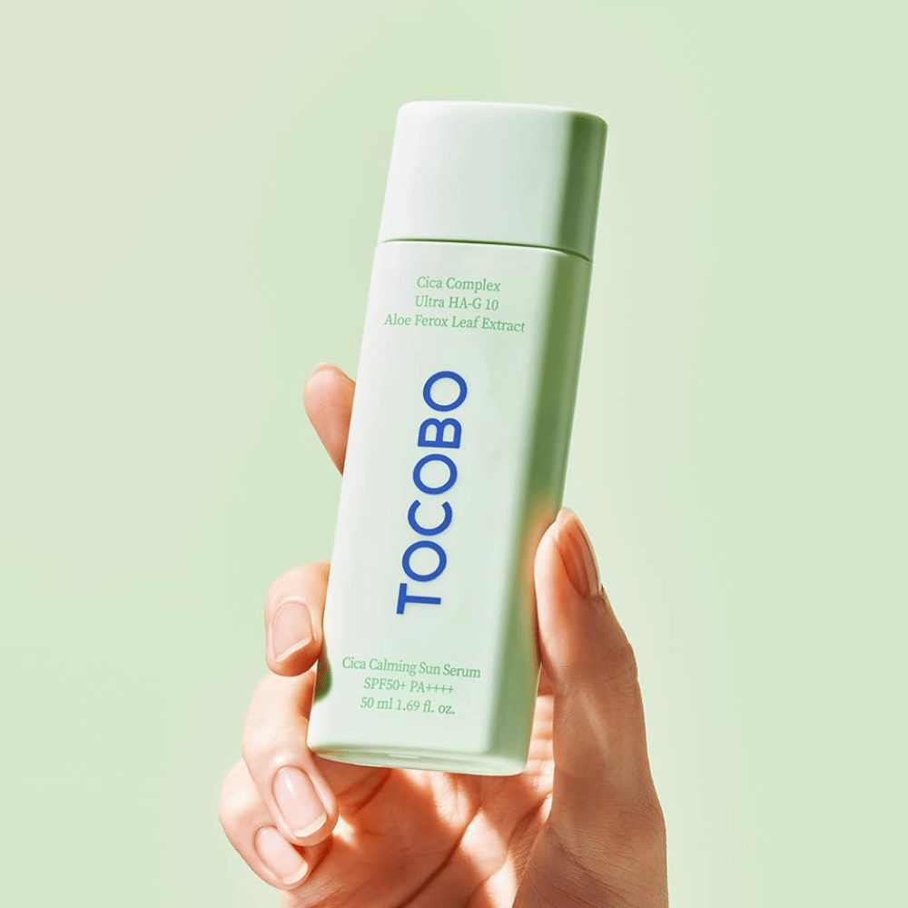 Mano sosteniendo TOCOBO Cica Calming Sun Serum con empaque verde menta y texto azul contra un fondo verde claro.