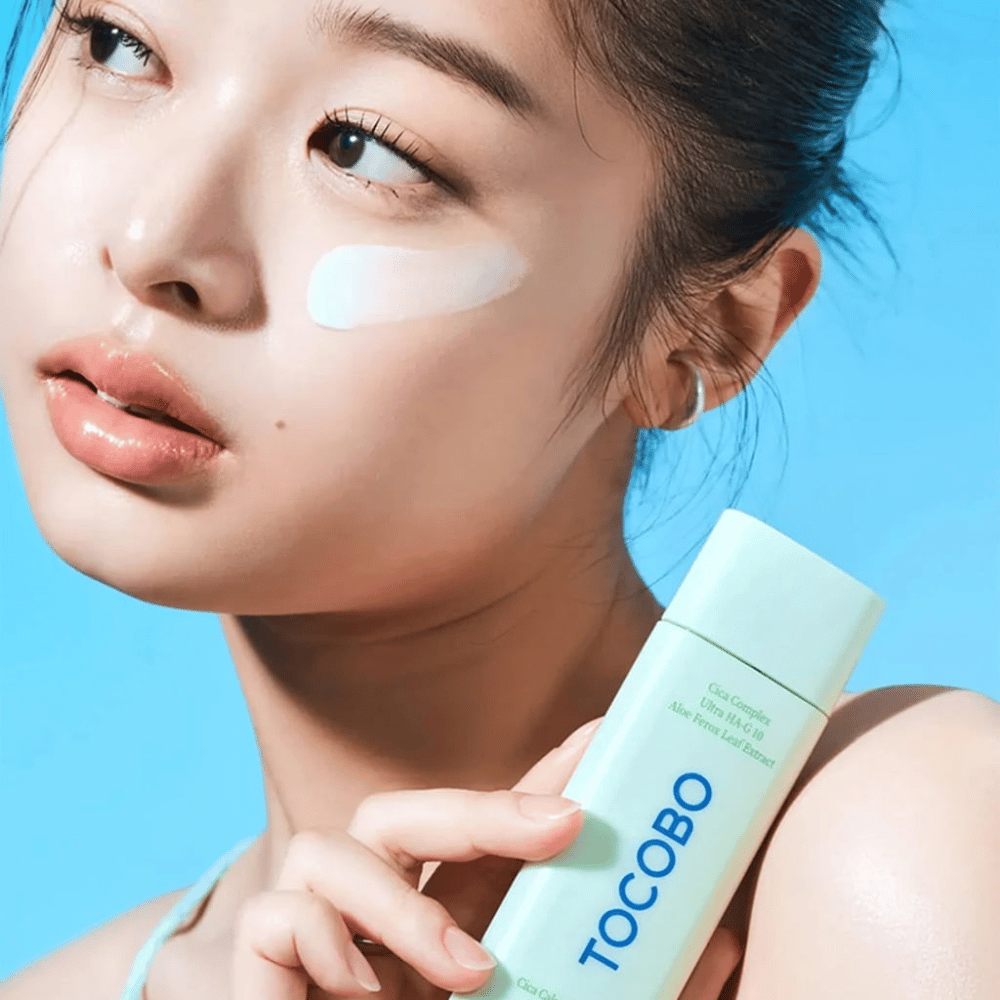 Modelo con protector solar bajo el ojo sosteniendo TOCOBO Cica Calming Sun Serum en empaque verde menta contra un fondo azul.