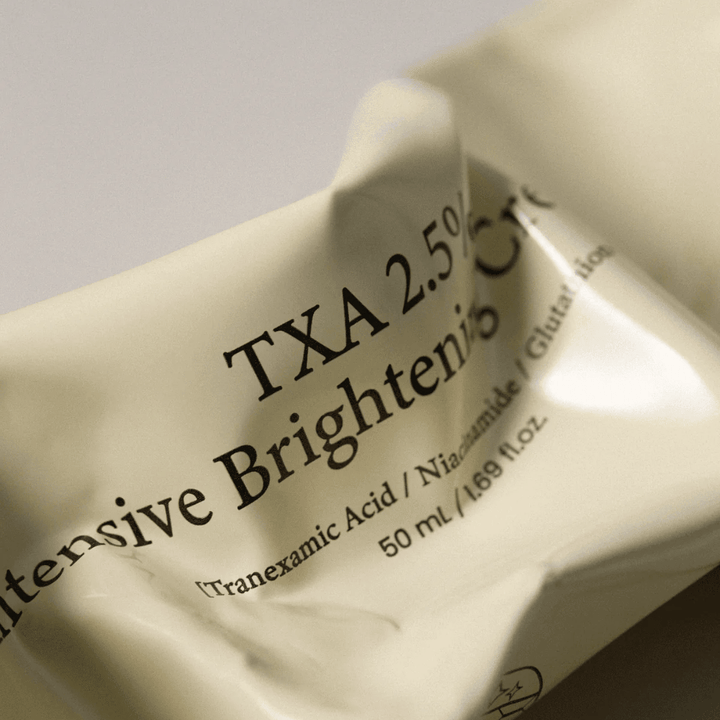 Primer plano de AXIS - Y TXA 2.5% Intensive Brightening Cream donde el diseño suave y minimalista del tubo es el centro de atención. La imagen destaca el nombre del producto, Tranexamic Acid y Niacinamide, así como el elegante envase beige.