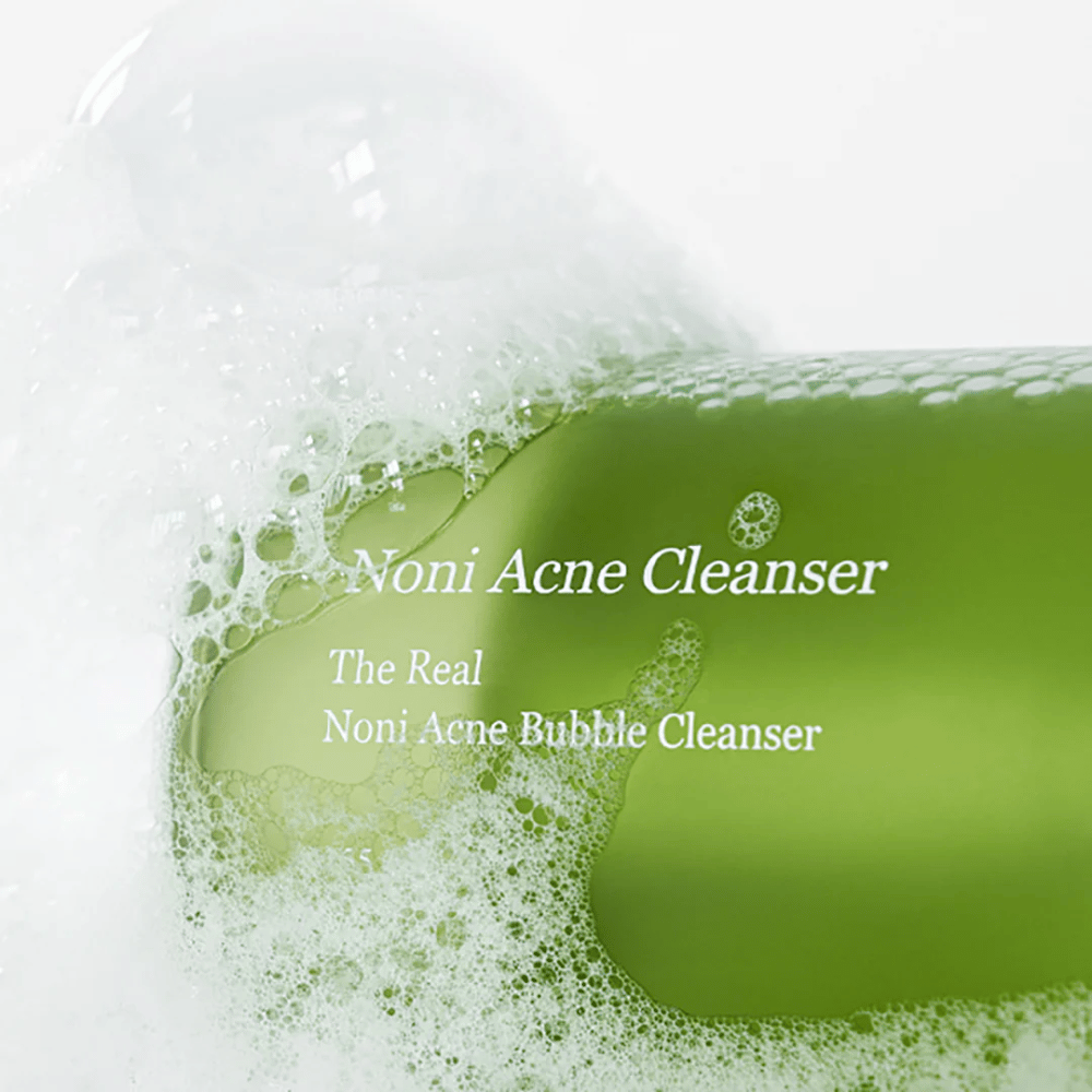 La botella con The Real Noni Acne Bubble Cleanser está en foco y rodeada de abundante espuma. El color verde fresco señala ingredientes naturales, perfecto para limpiar y combatir el acné. Un producto ideal para una piel limpia y clara.