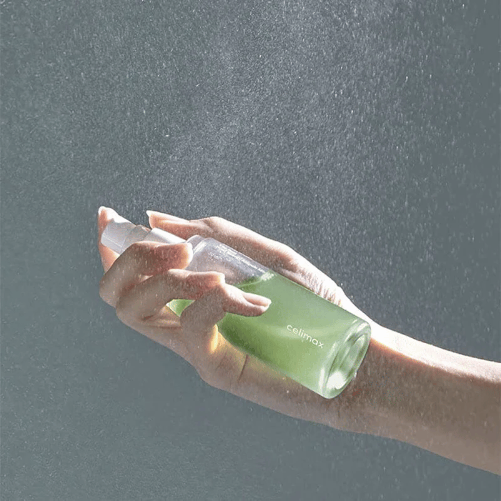 Una mano sostiene Celimax Noni Mist botella de spray y aplica el producto en una fina y uniforme pulverización sobre la piel. La botella es verde y el producto se comercializa como "The Real Noni Energy Ampule Mist" para proporcionar energía e hidratación a la piel.