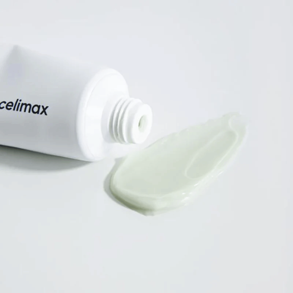 Celimax Noni Energy Repair Cream en un tubo blanco con una pequeña cantidad de crema verde claro esparcida al lado del tubo sobre una superficie blanca. La crema está destinada a nutrir y reparar la piel.