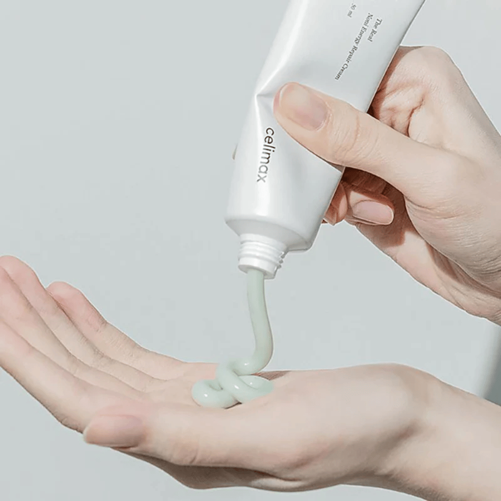 Una mano sostiene un tubo blanco con Celimax Noni Energy Repair Cream, y la crema se exprime en un chorro uniforme sobre la palma de la mano. La crema es verde claro y se comercializa para nutrir y restaurar la piel.