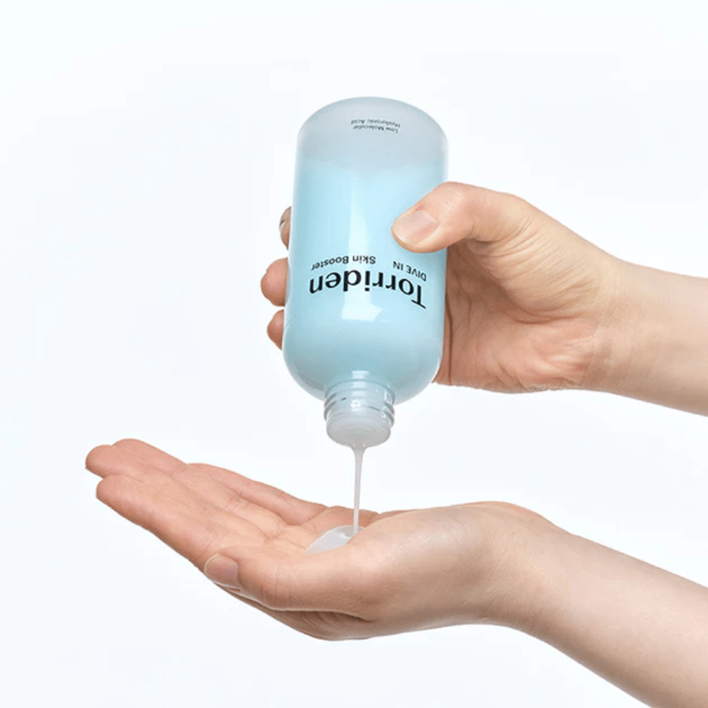 Manos que sostienen una botella de producto para el cuidado de la piel y dosifican un líquido claro en la palma. La botella está etiquetada con "Torriden - DIVE-IN Low Molecule Hyaluronic Acid Skin Booster".