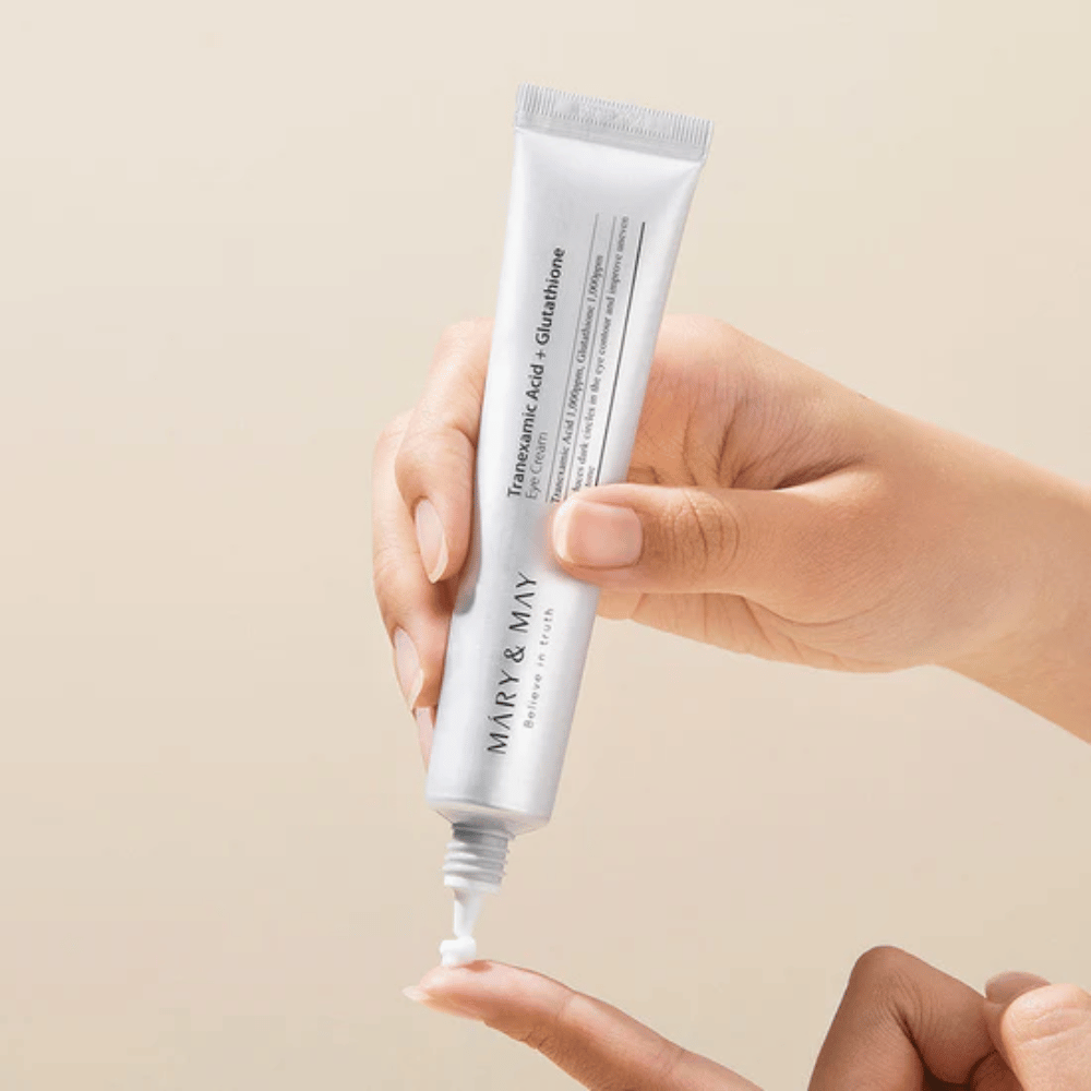 Una mano sostiene un tubo de MARY & MAY "Tranexamic Acid + Glutathione Eye Cream". El tubo es blanco y el diseño es limpio y sencillo, lo que resalta las propiedades calmantes e iluminadoras del producto para el área de los ojos. El producto se aplica suavemente, con un enfoque en la precisión y la eficiencia.