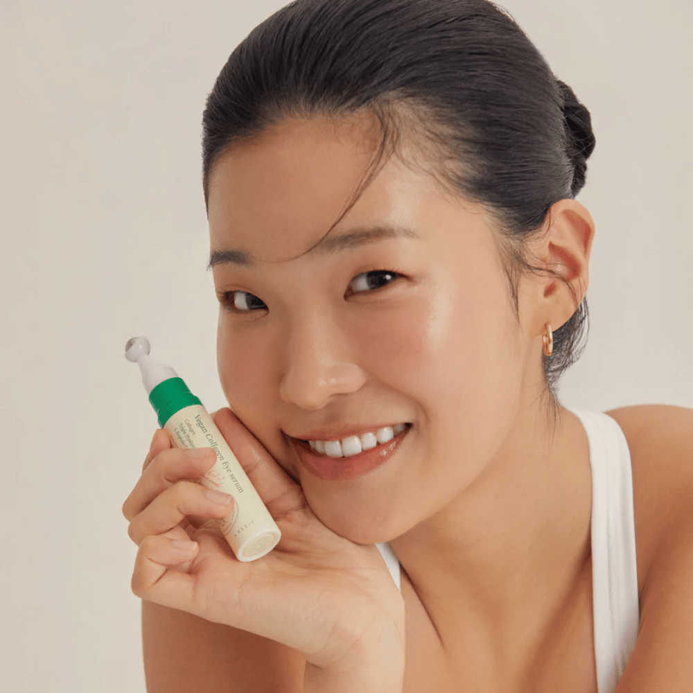 Una mujer con el cabello recogido y pendientes dorados sostiene un AXIS-Y Vegan Collagen Eye Serum 10ml cerca de su rostro, mientras sonríe suavemente. El suero está diseñado para hidratar y reducir arrugas alrededor de los ojos, especialmente formulado para piel sensible.