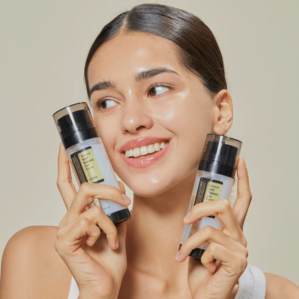 Mujer sonriente con piel brillante sostiene dos botellas COSRX Advanced Snail Radiance Dual Essence 80 ml cerca de la cara contra un fondo claro.