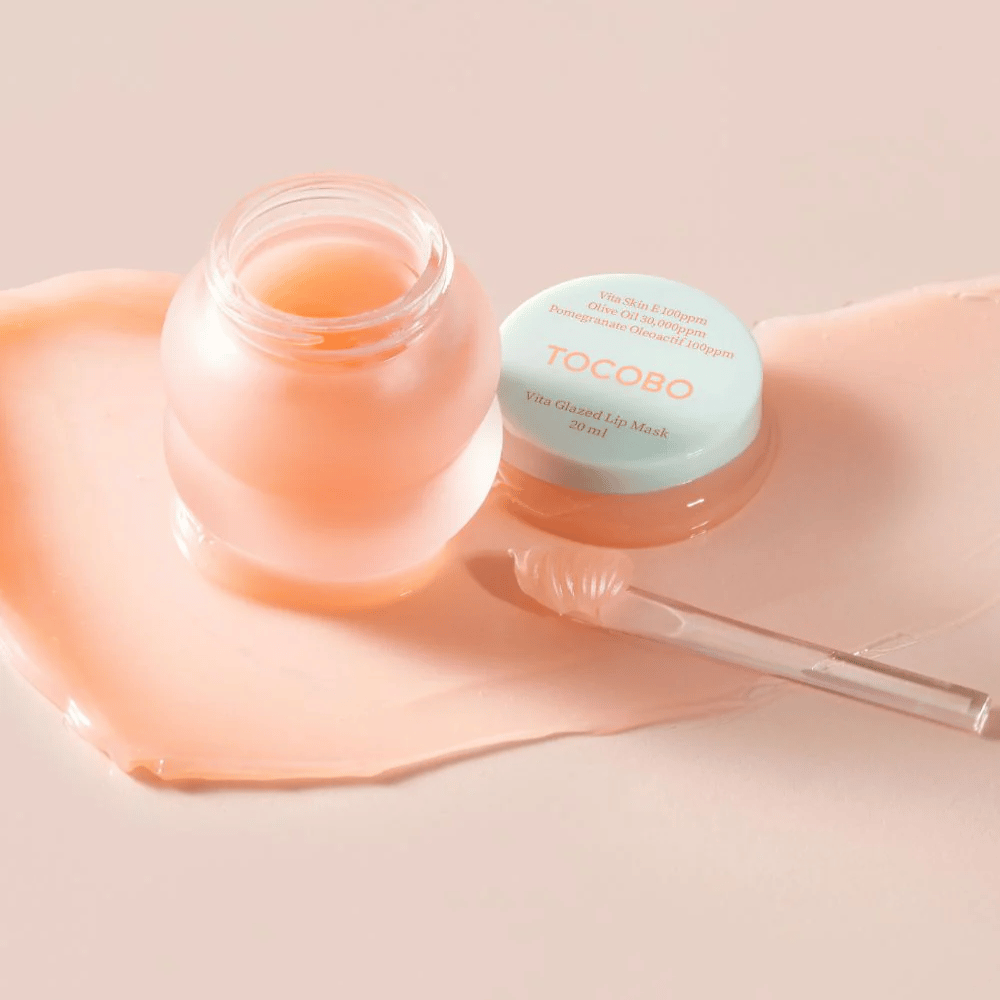 Un frasco de "Vita Glazed Lip Mask" de Tocobo se muestra. El frasco es transparente con una tapa turquesa clara y contiene una mascarilla labial brillante y rosa. Una pequeña espátula está al lado del frasco, y parte de la mascarilla está esparcida sobre la superficie. El producto está diseñado para hidratar y cuidar los labios.