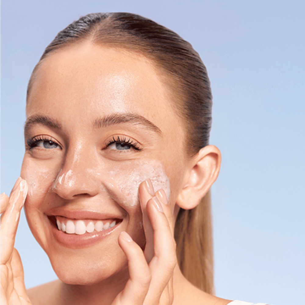Mujer sonriente aplica LANEIGE Water Bank Gentle Gel Cleanser en forma espumosa en las mejillas, sobre fondo azul.