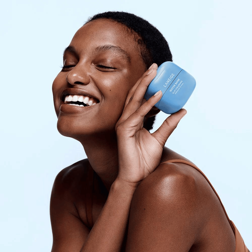 Mujer sonriente sostiene un frasco azul de LANEIGE Water Bank Blue Hyaluronic Cream contra su mejilla, con fondo azul claro.