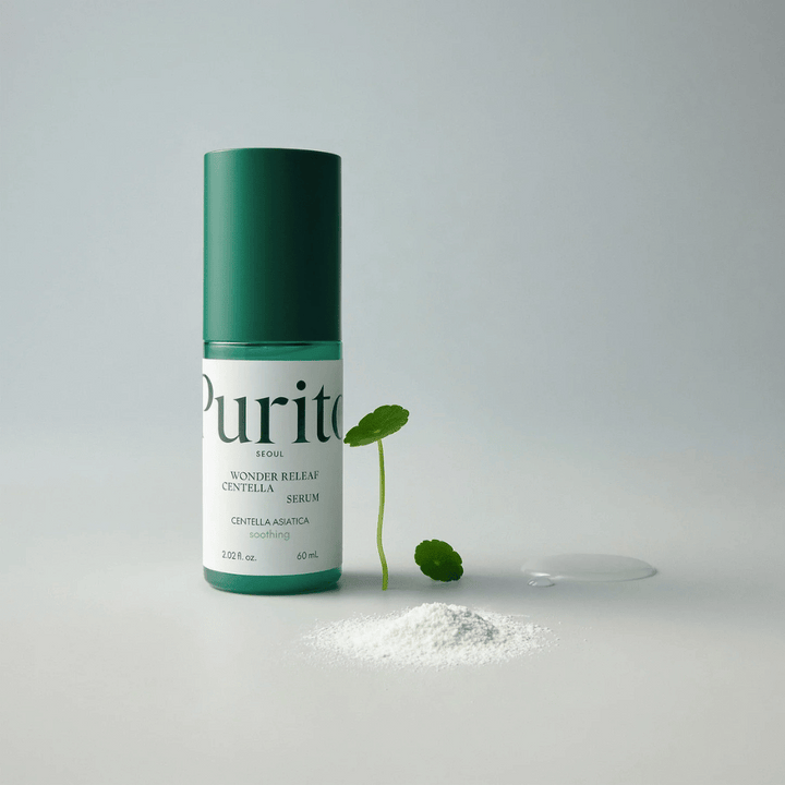 Grön flaska av Purito Wonder Releaf Centella Serum 60 ml står på ett vitt underlag, omgiven av centellablad, ett genomskinligt serumdropp och ett litet vitt pulver.