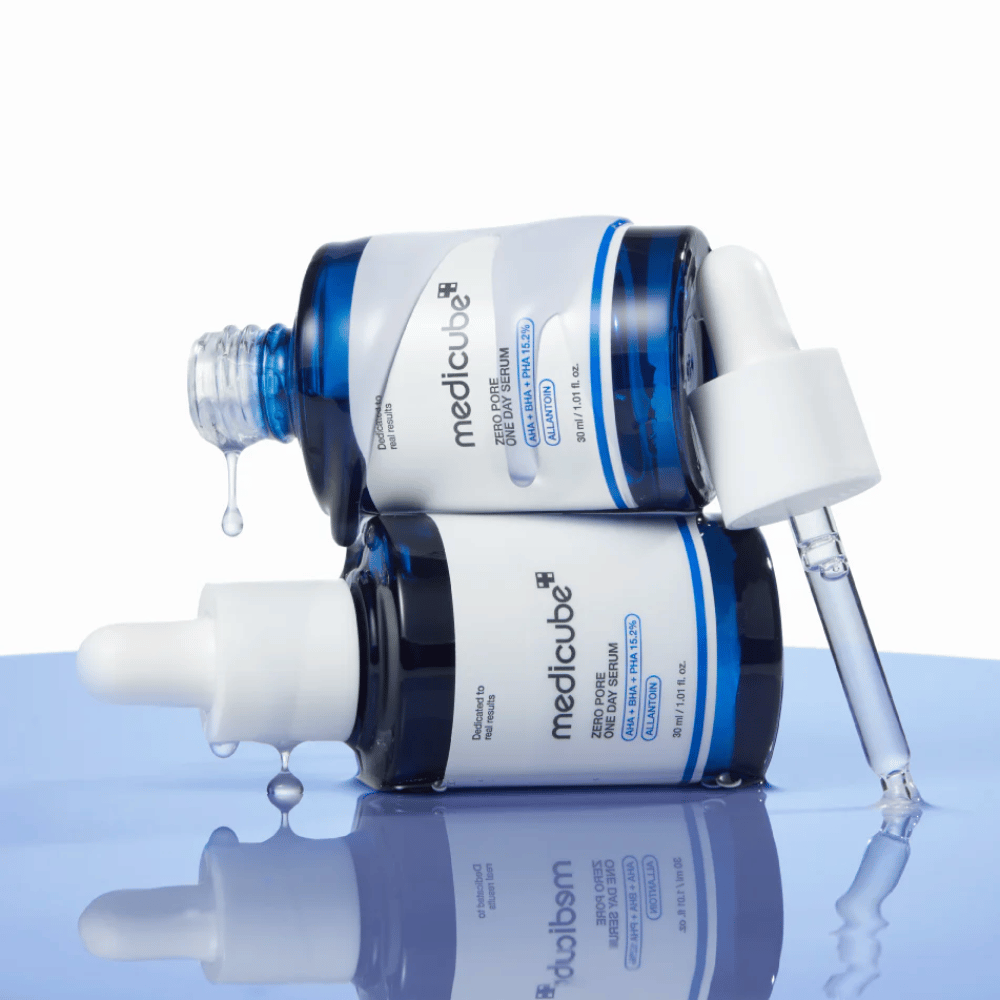 Dos botellas azules de Medicube Zero Pore One Day Serum acostadas una contra la otra, con pipetas y gotas de suero sobre una superficie azul reflectante.