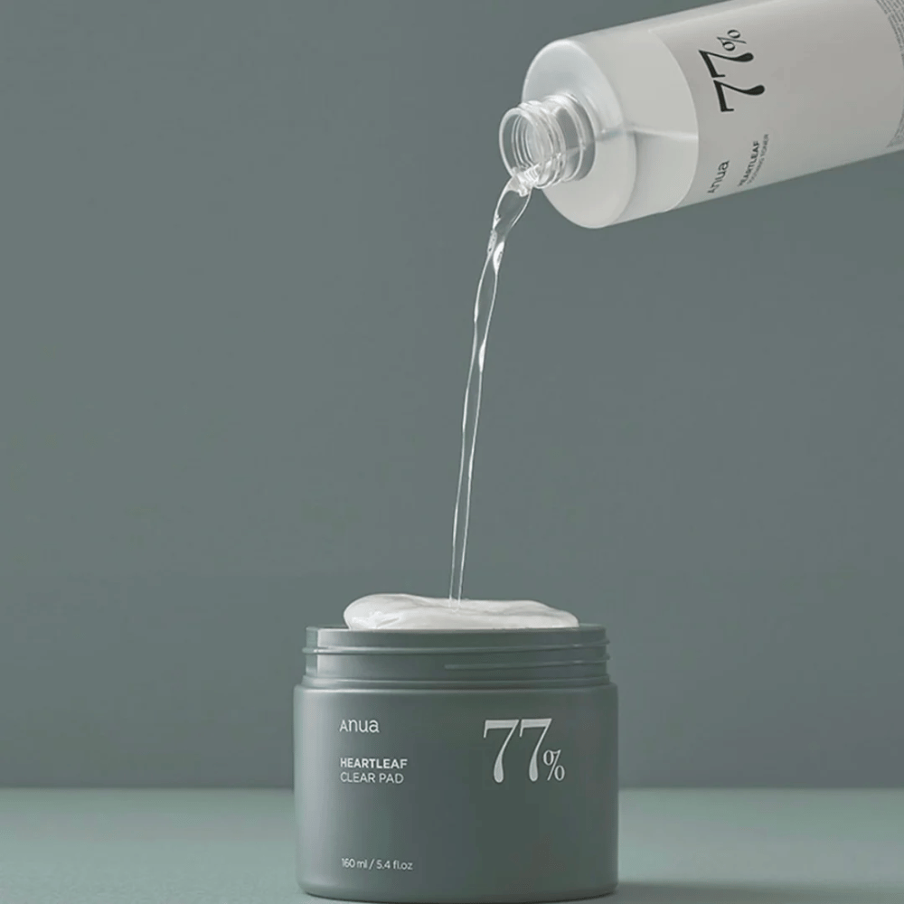 Una botella transparente vierte un líquido en un frasco gris etiquetado "anua HEARTLEAF 77% Clear Pad", que contiene una pila de discos de cuidado de la piel blancos. El fondo es simple y de un solo color en un suave tono gris.