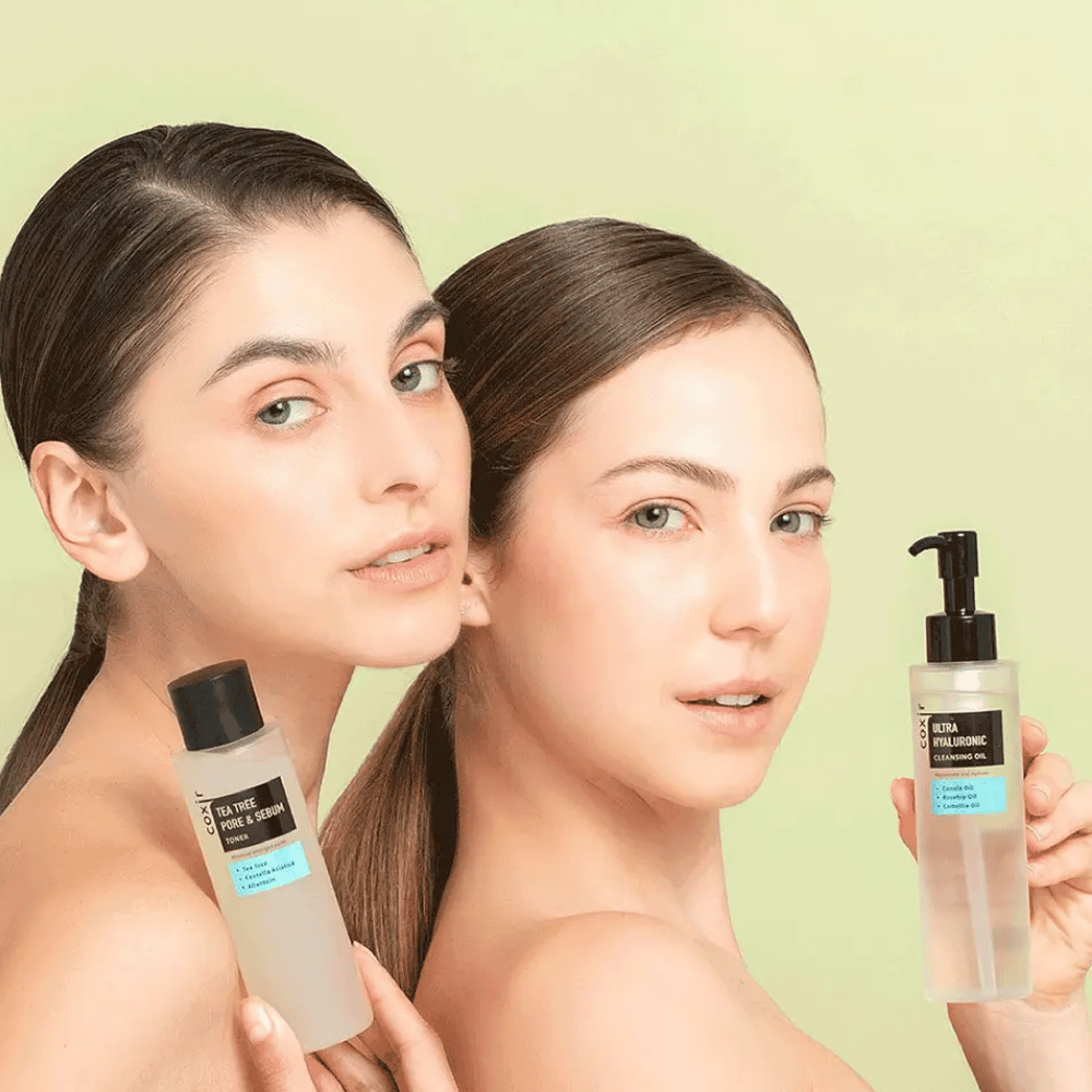 Dos mujeres con hombros descubiertos posan con productos para el cuidado de la piel; una sostiene una botella etiquetada "TEA TREE PORE & SEBUM", y la otra una botella de bomba etiquetada "ULTRA HYALURONIC CLEANSING OIL". Están frente a un fondo verde claro.
