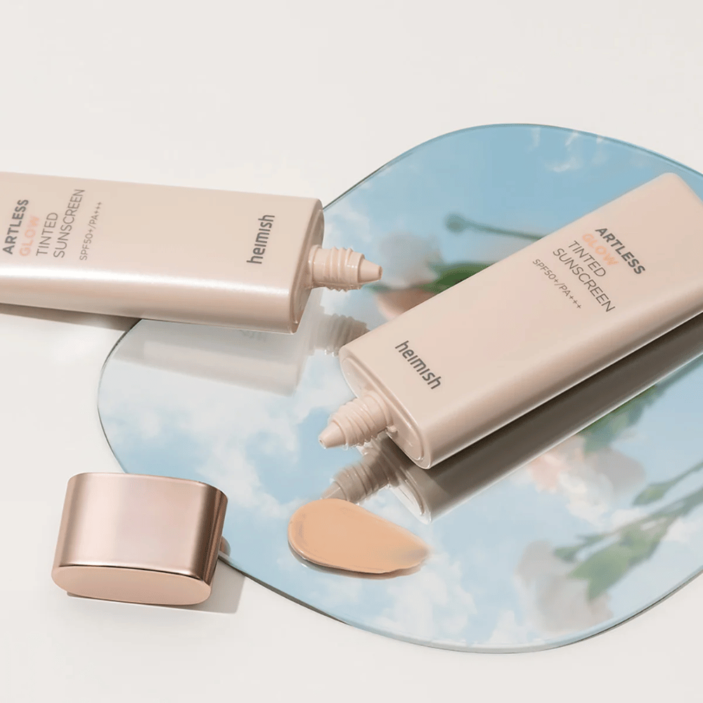 Una botella de heimish Artless Glow Tinted Sunscreen SPF50+ PA+++ 40ml y una botella de concealer sobre un espejo, que muestra diferentes tonos de piel.