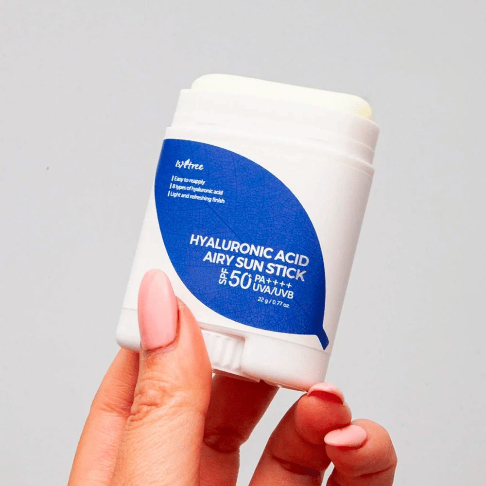 Una mano sostiene un protector solar blanco con etiqueta azul de la marca Isntree. El producto se llama "Hyaluronic Acid Airy Sun Stick" y tiene un factor de protección solar SPF 50+ PA++++.