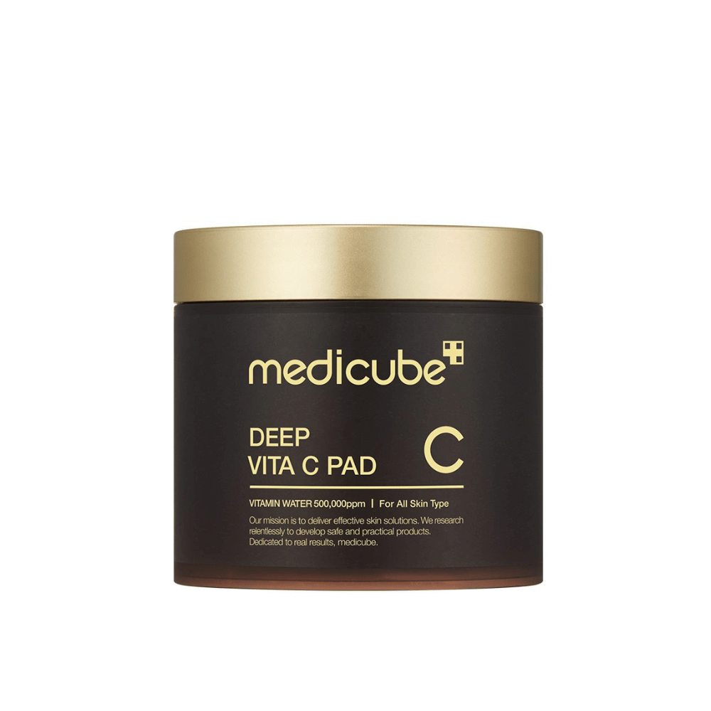 Un tarro de medicube, marcado "Deep Vita C Pad", con tapa dorada y cuerpo oscuro. El texto en el tarro indica vitamina C y es apto para todo tipo de piel. El fondo es neutro.
