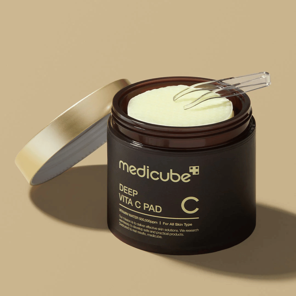Un tarro con medicube Deep Vita C Pad. El tarro es marrón oscuro con texto dorado y una etiqueta blanca invertida con información sobre vitamina C. El interior de la tapa es dorado, y unas pinzas transparentes descansan sobre los discos de algodón amarillo claro en su interior. El fondo es de color beige sólido.