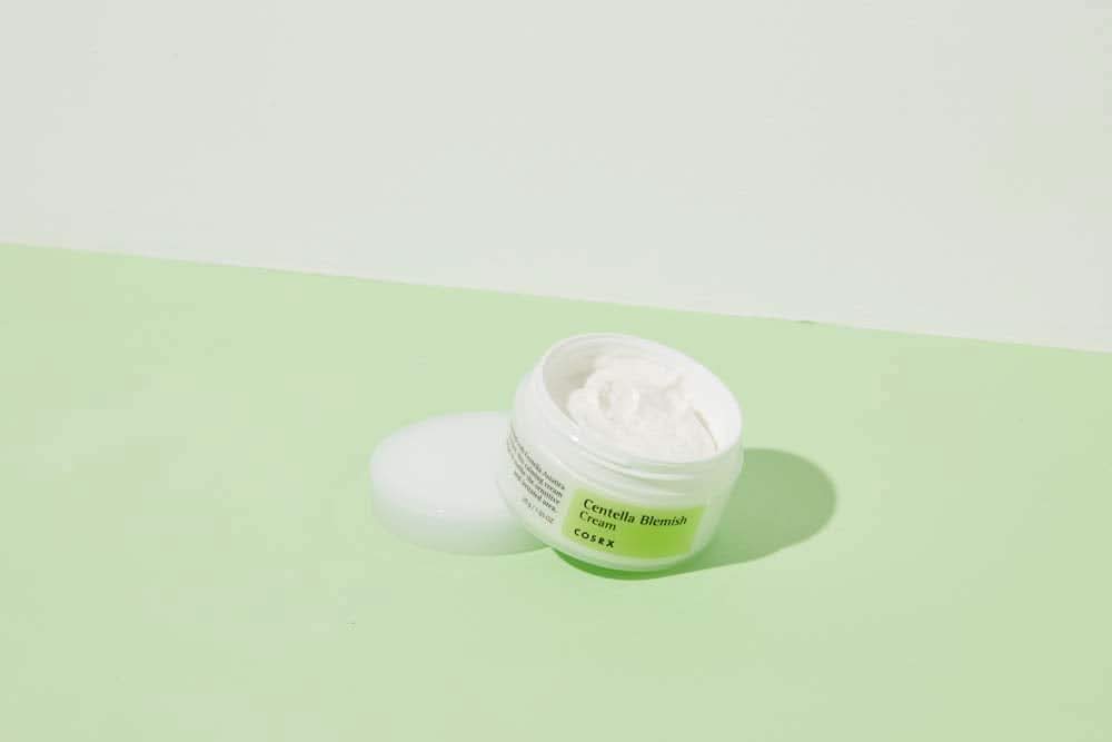 Un frasco COSRX Centella Blemish Cream 30ml que está sobre una superficie verde.