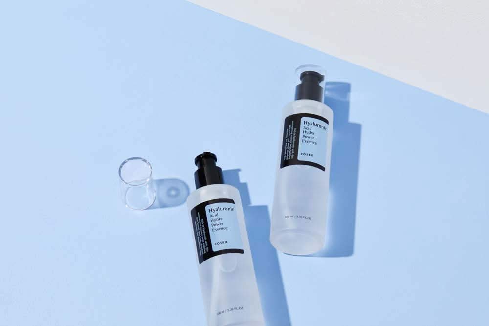 El COSRX:s Hyaluronic Acid Hydra Power Essence de 100 ml proporciona beneficios hidratantes sobre la superficie azul.