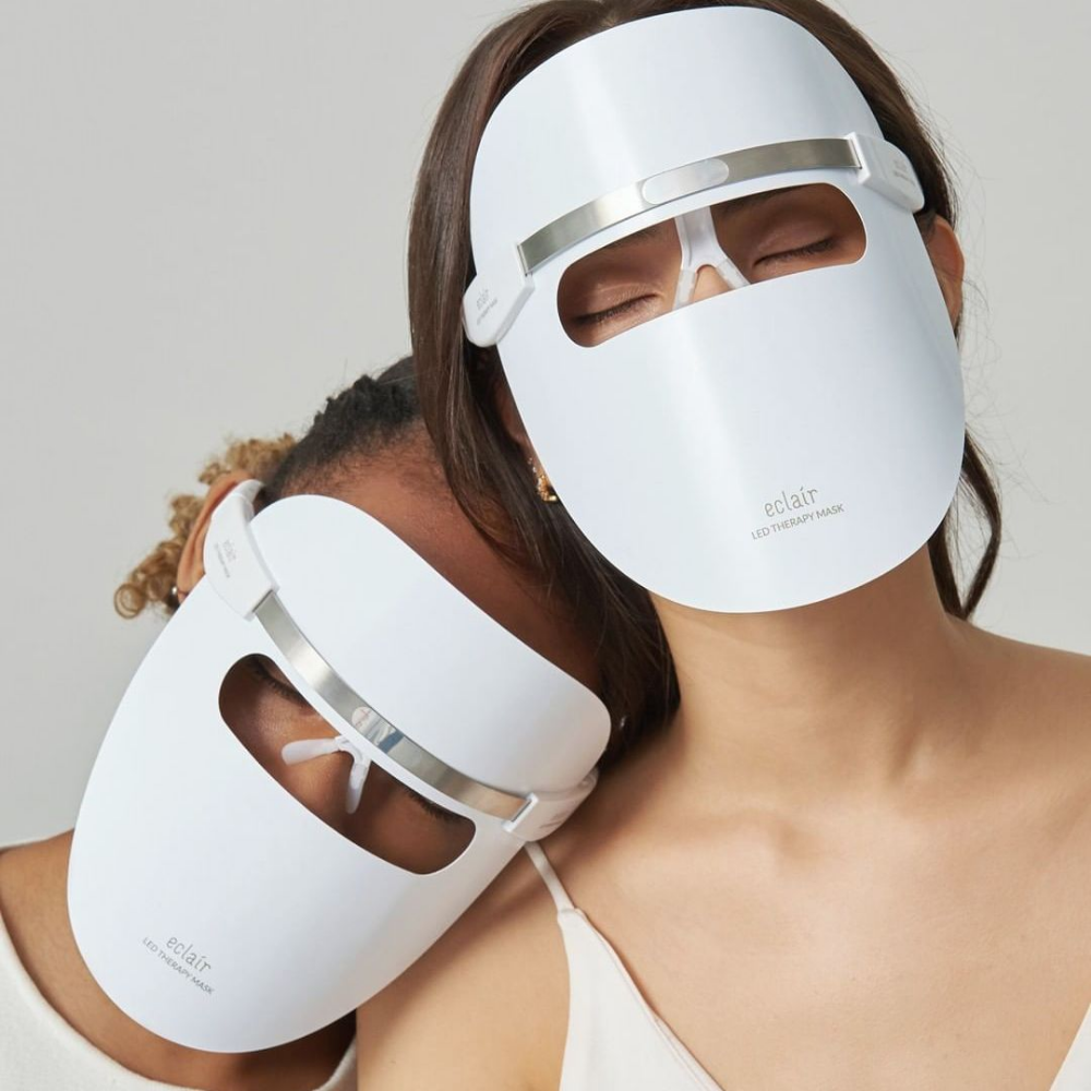 Dos mujeres que llevan máscaras blancas demuestran ECLAIR LED Therapy Mask para mejoras del tono de piel.