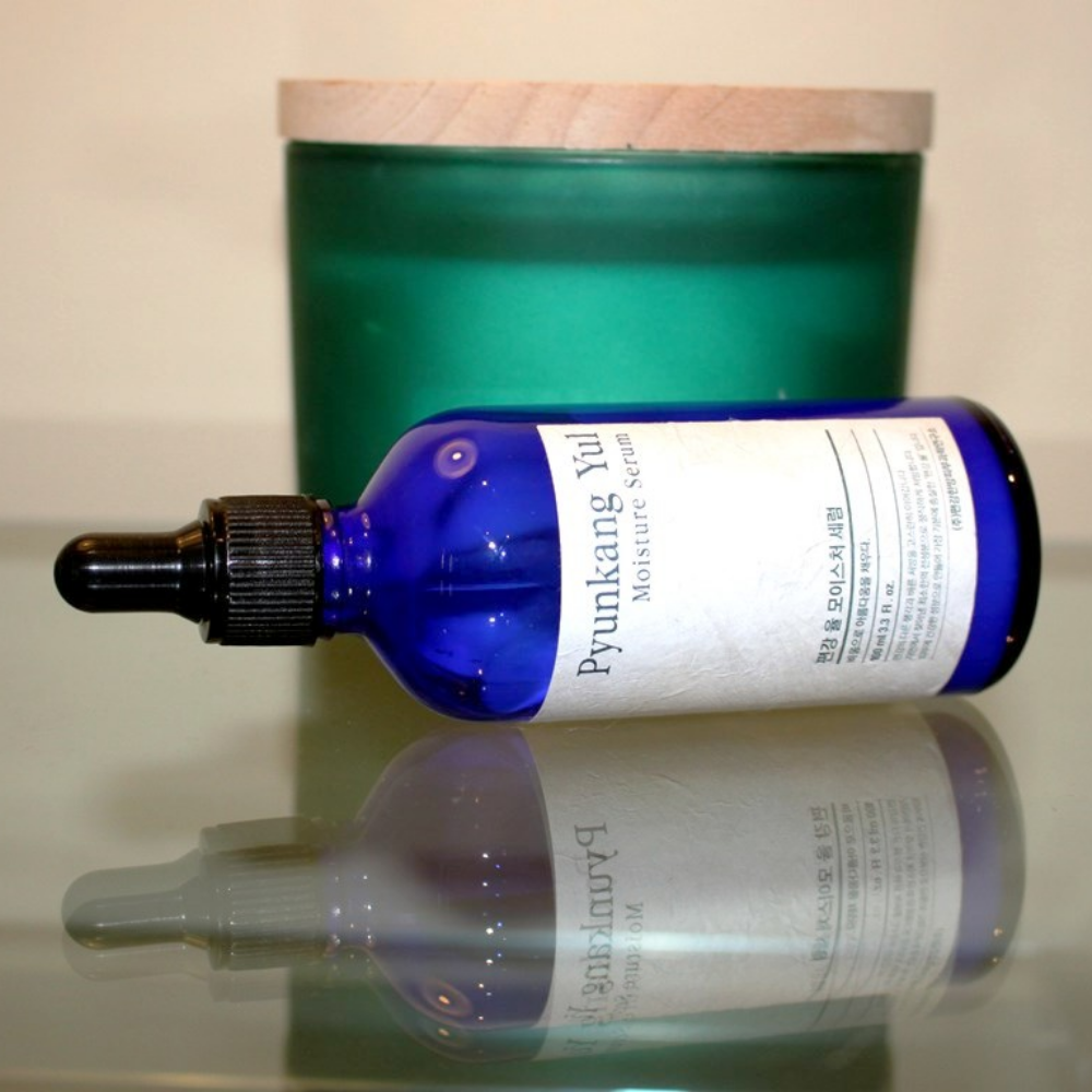 Una botella Pyunkang Yul Moisture Serum 100ml equilibra los niveles de humedad en una mesa al lado de una vela.