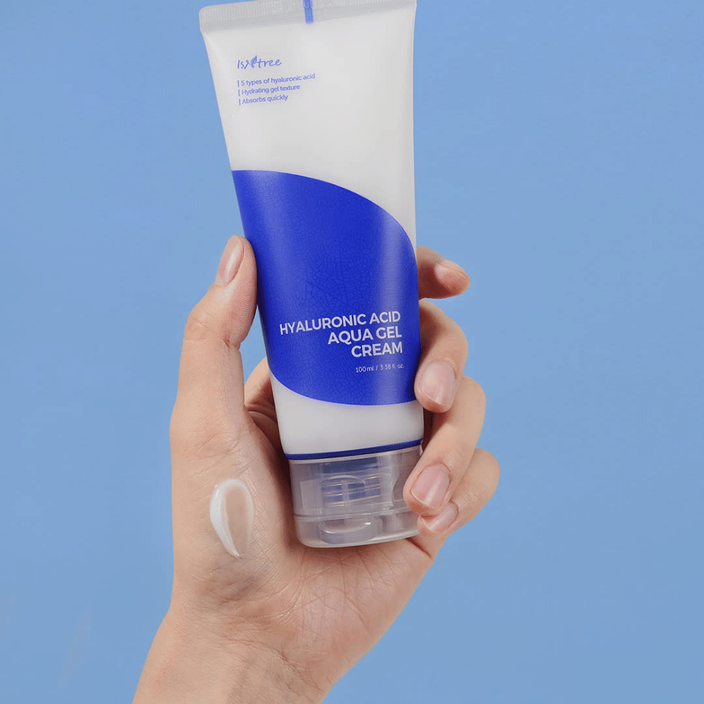 Una mano sosteniendo un tubo Isntree Hyaluronic Acid Aqua Gel Cream de 100ml sobre un fondo azul.